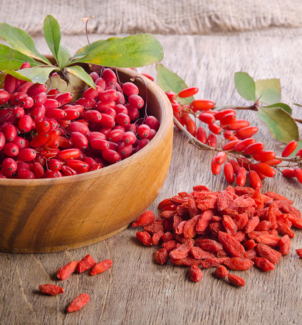 a wooden bowl filled with clusters of Goji berries and dried goji berries next to the bowl on a wooden table