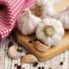 Four garlic bulbs and a few cloves on a wood cutting board, on a white washed wood table with a red and white gingham towel